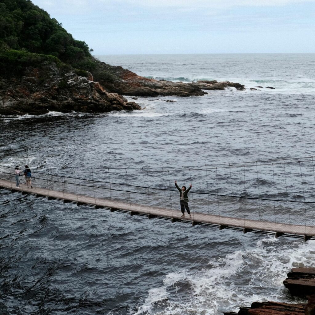 A scenic suspension bridge with people crossing above turbulent ocean waves.